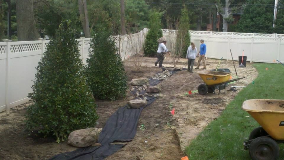 Three people work in a residential backyard, planting trees and installing landscaping fabric near a white vinyl fence.