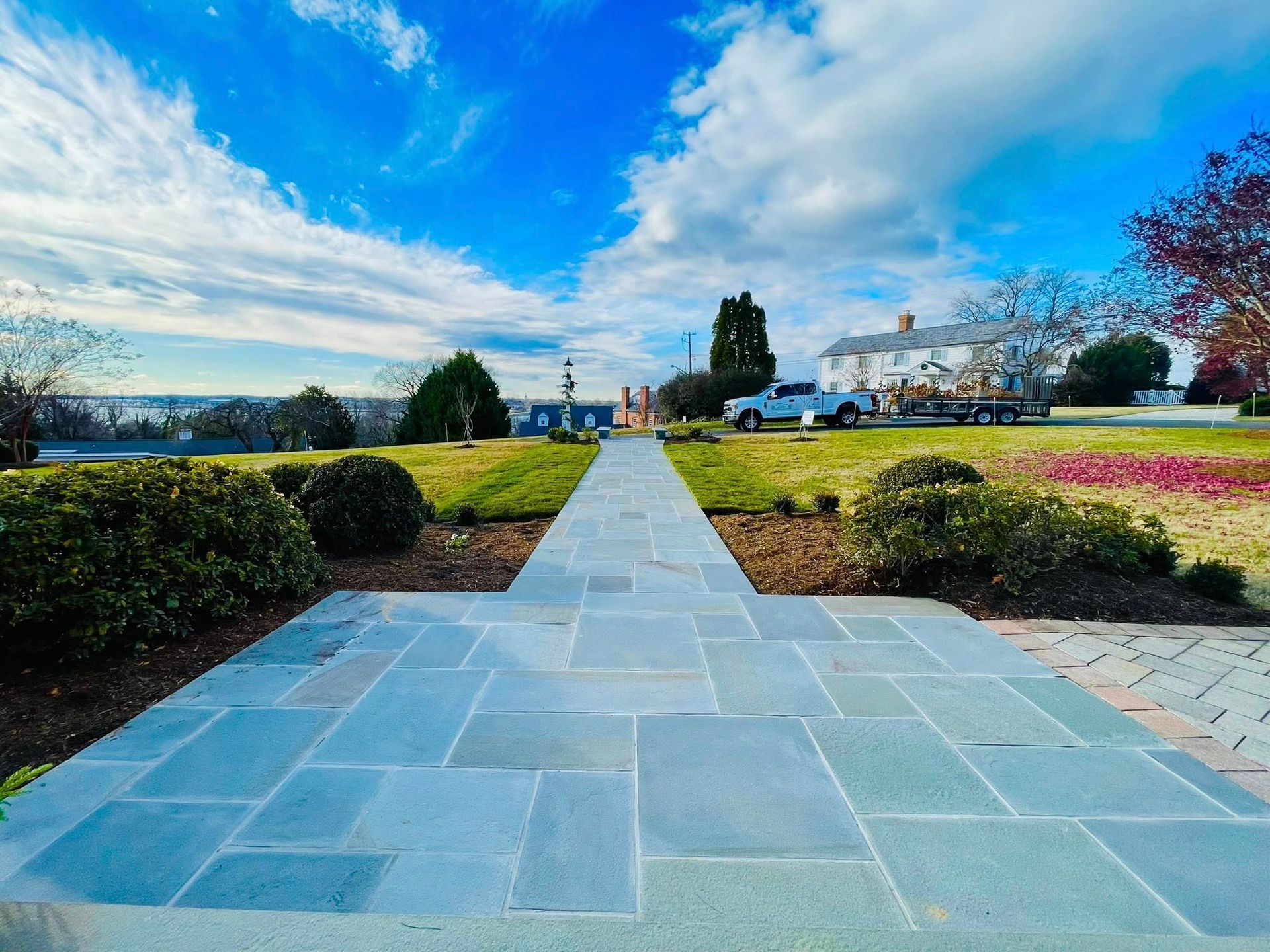 A stone pathway leads through a grassy yard toward a house under a bright blue, cloudy sky.
