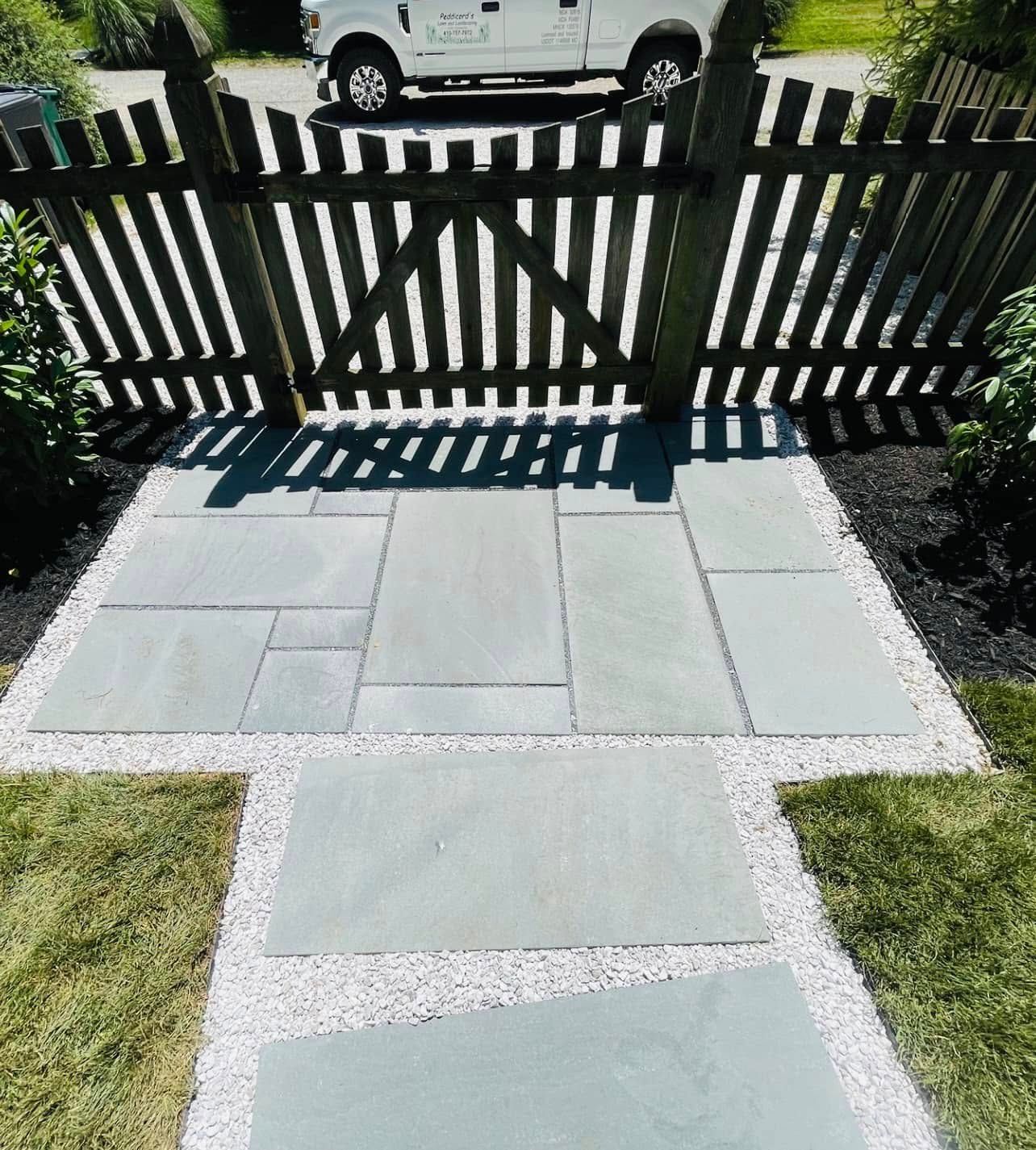 A gray stone patio and walkway set in white gravel leading to a wooden fence with a white vehicle parked behind it.
