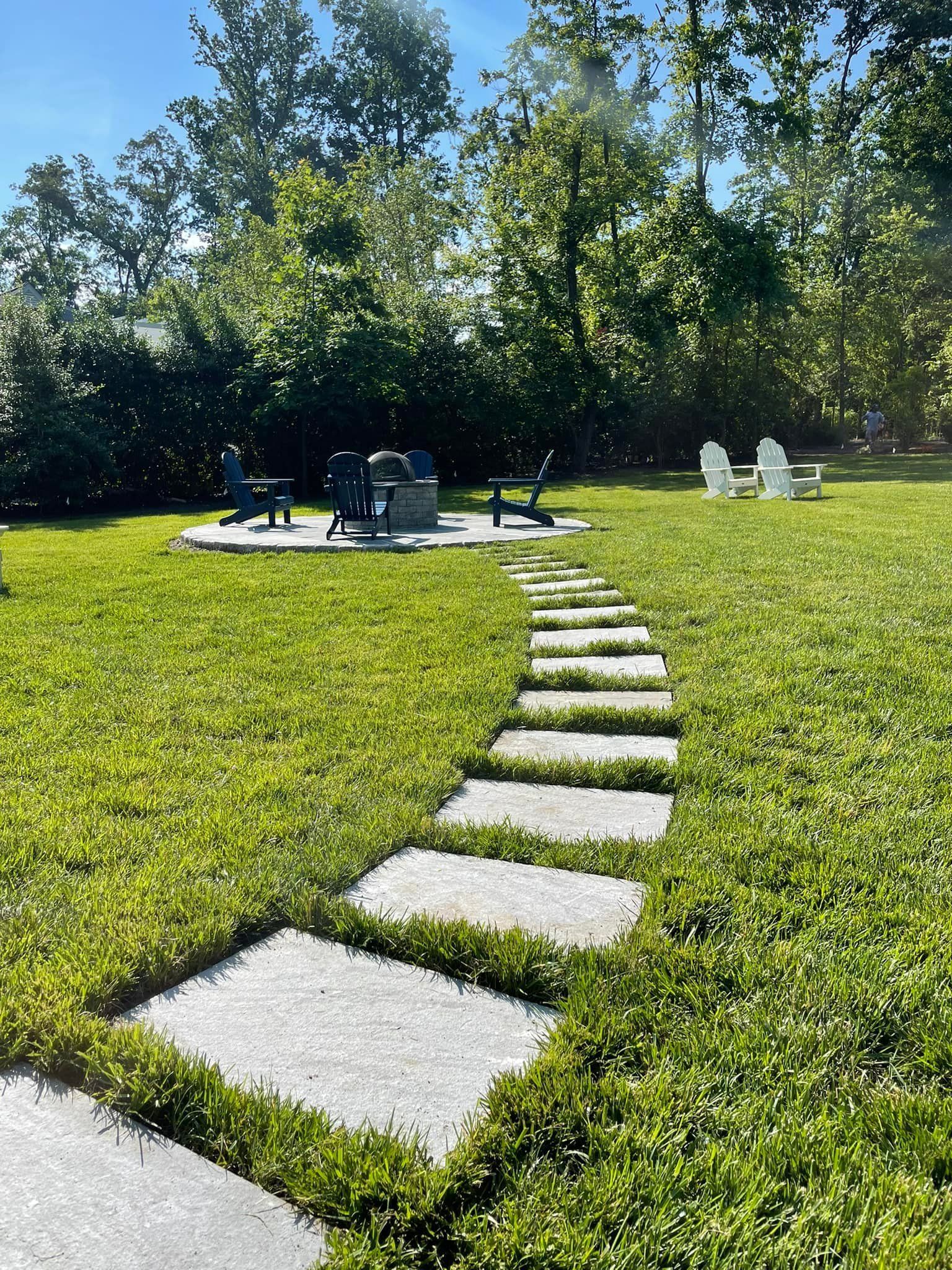 A stone pathway leads across a green lawn to a fire pit area with chairs surrounded by trees under a clear blue sky.