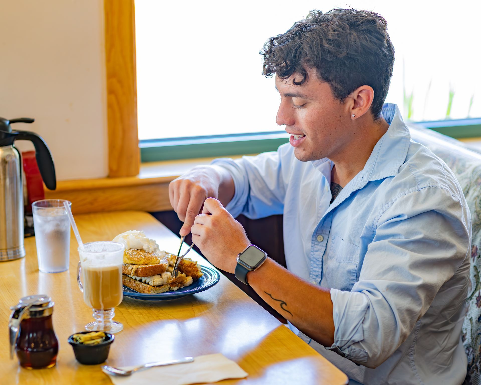 Person eating breakfast at a table in a restaurant. He has a blue shirt and is using a fork and knife.