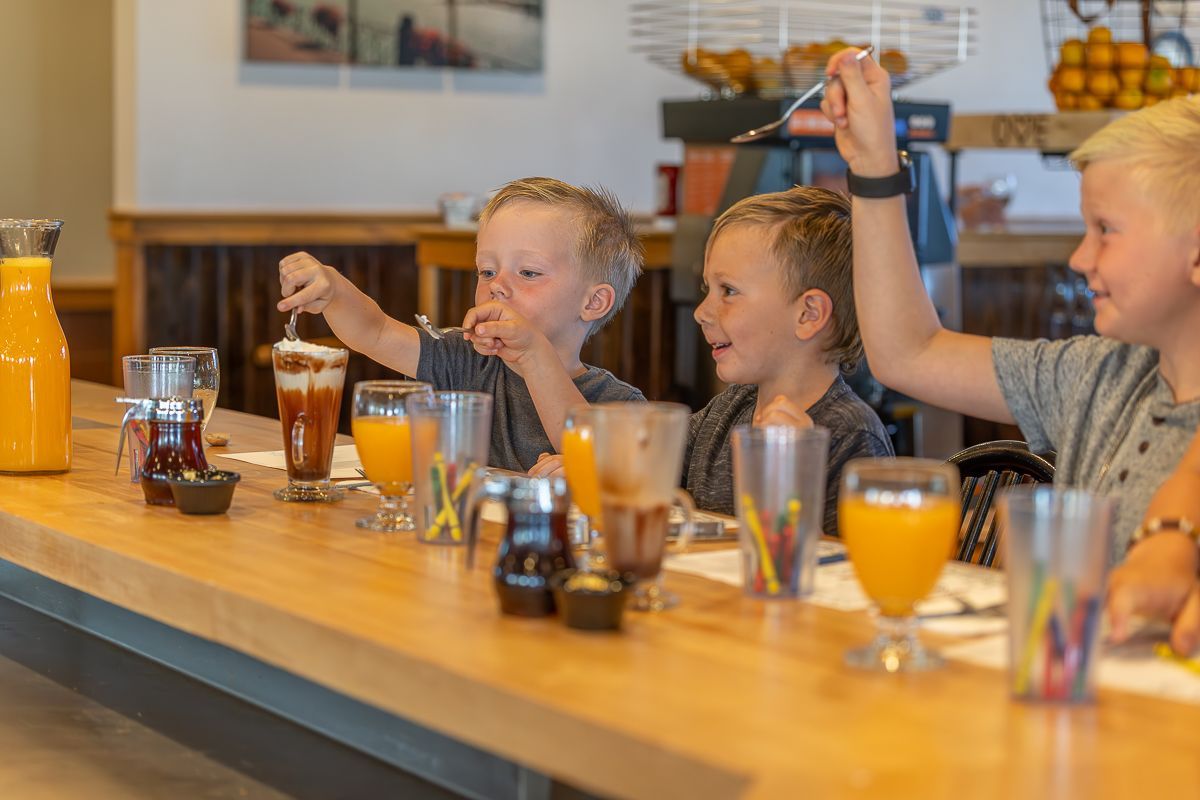 Three children at a counter eating and drinking beverages, holding forks. Orange juice in glasses.