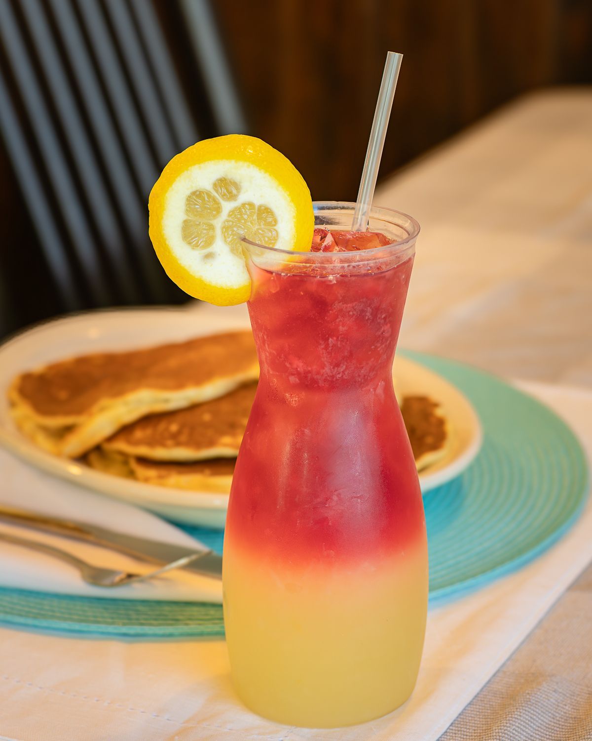 A layered drink with lemon slice and straw, next to pancakes on a turquoise plate.