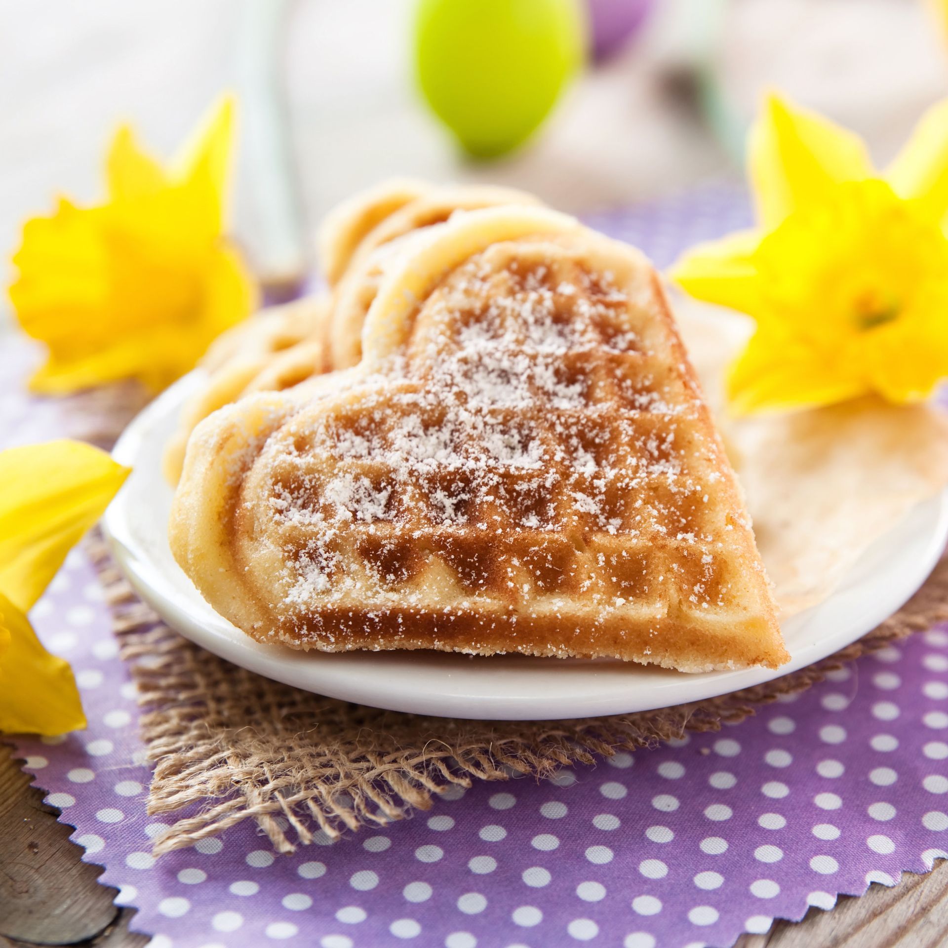 Heart-shaped waffles dusted with powdered sugar on a white plate with yellow flowers and a purple polka-dot cloth