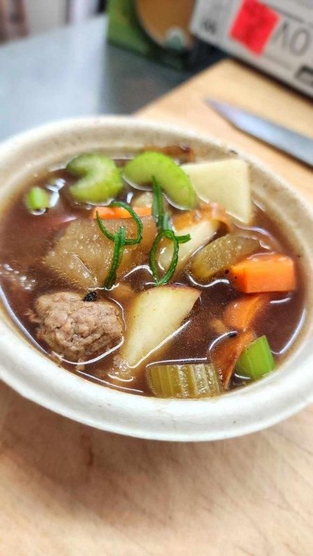 A bowl of soup with meat, potatoes, carrots and celery on a wooden table.