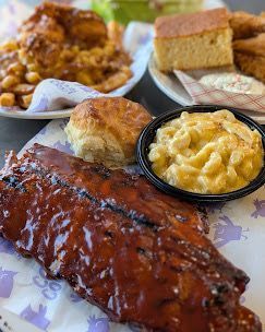 Platters of BBQ ribs, mac & cheese, biscuit, and chicken with sides on patterned paper.
