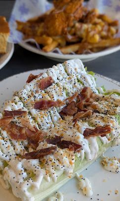 Wedge salad with bacon and dressing on a white plate, fries and chicken in background.