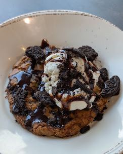 Cookie dessert with ice cream, Oreo pieces, and chocolate sauce in a white bowl.
