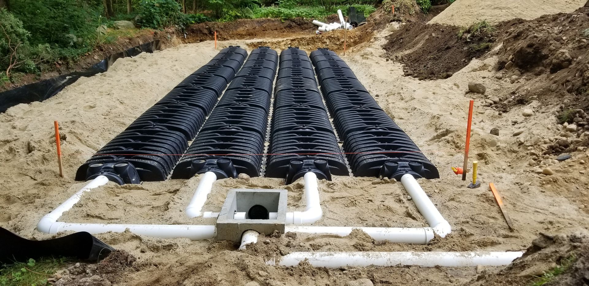 Construction site with four black septic tank chambers connected by white pipes, buried in sand.
