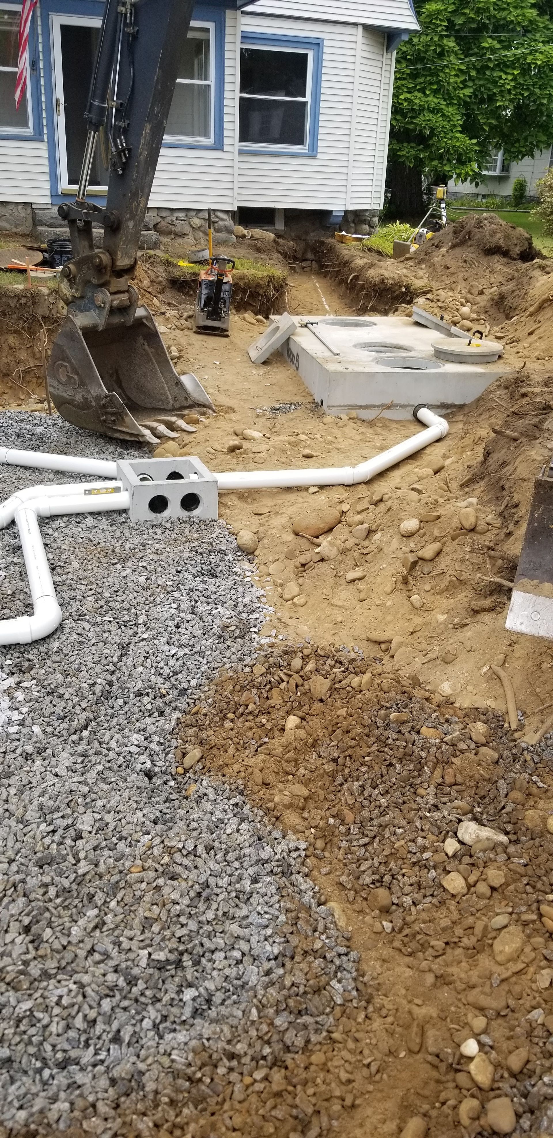 An excavator working on a construction project near a small house. Gravel and piping are visible in the excavation.