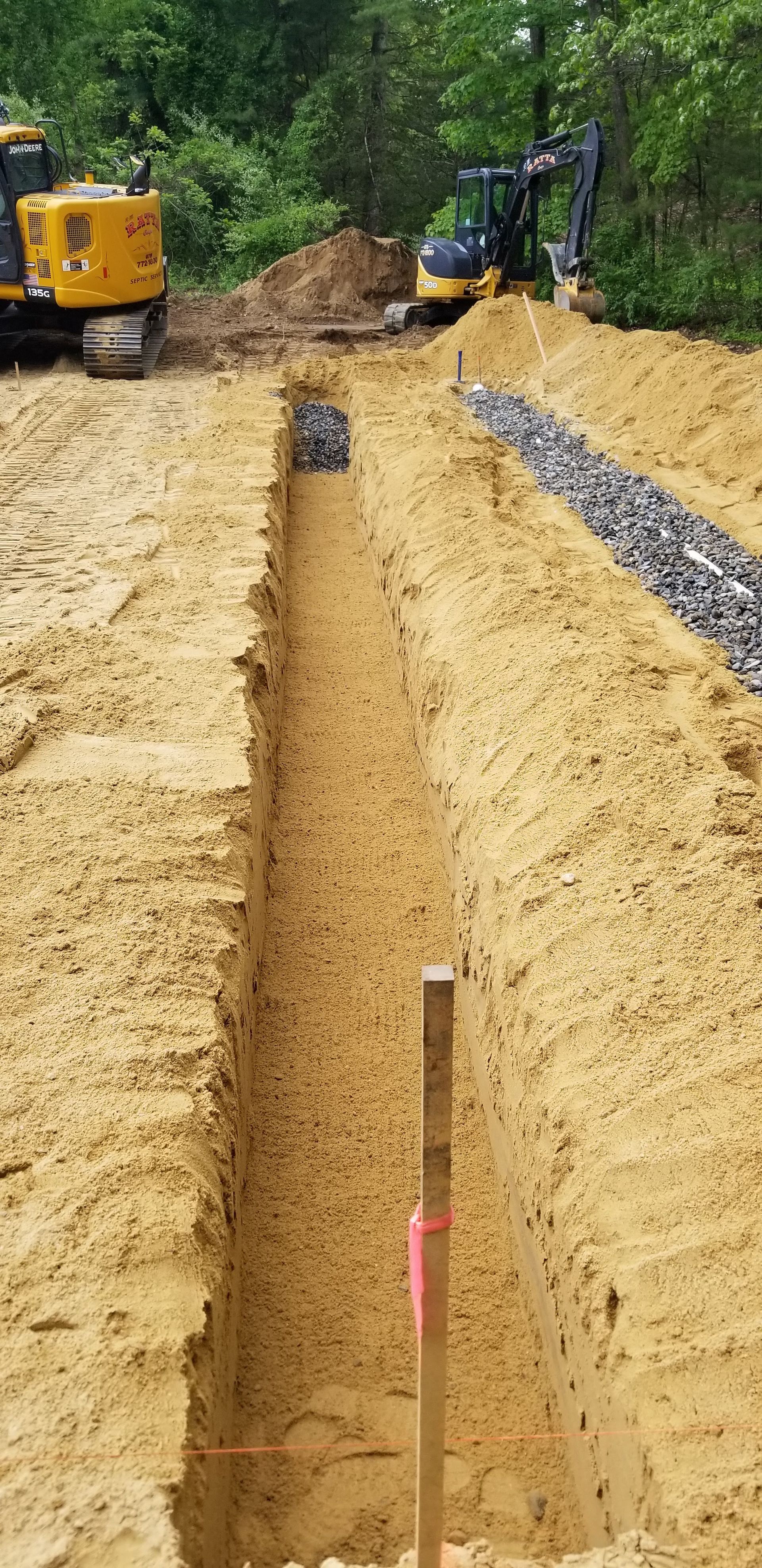 A long trench with gravel and an excavator in the background, likely for drainage. A wooden stake marks a point in the trench.