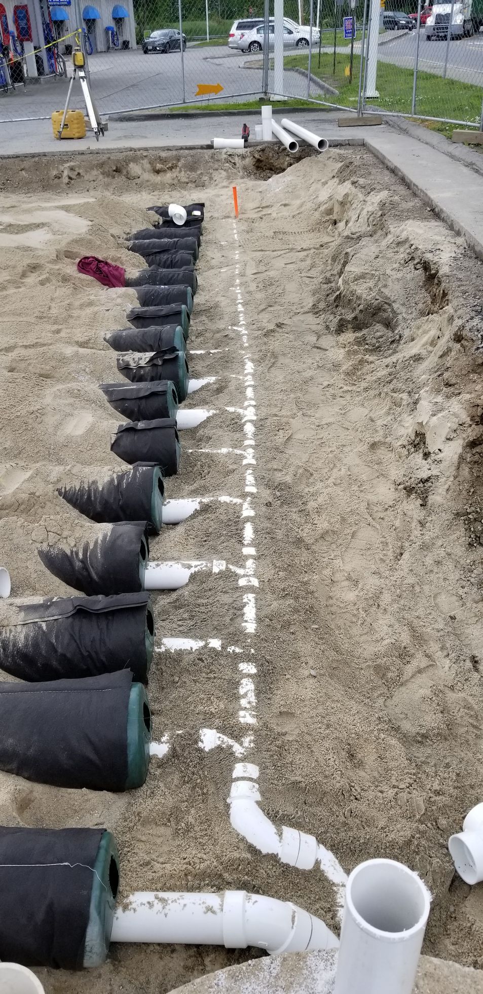 Construction site with a trench containing black cylindrical filters connected to white pipes, likely for drainage or water management.