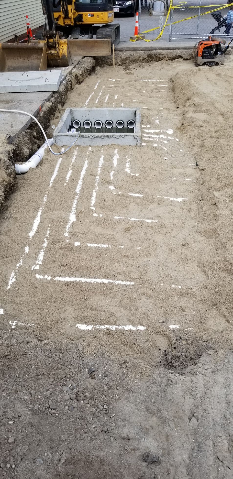 Construction site with a rectangular trench dug in the ground. White paint lines indicate the path for pipes. A backhoe and other equipment are visible.