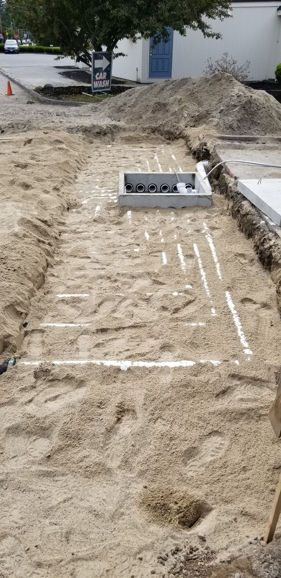A construction site with trenches in the sand, revealing white pipes. Concrete structures and a pile of dirt are in the background.