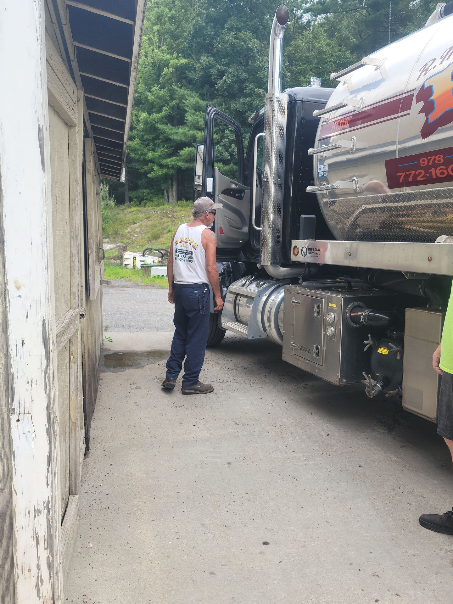Man standing next to a large tanker truck parked near a weathered building. The man wears a tank top and jeans.