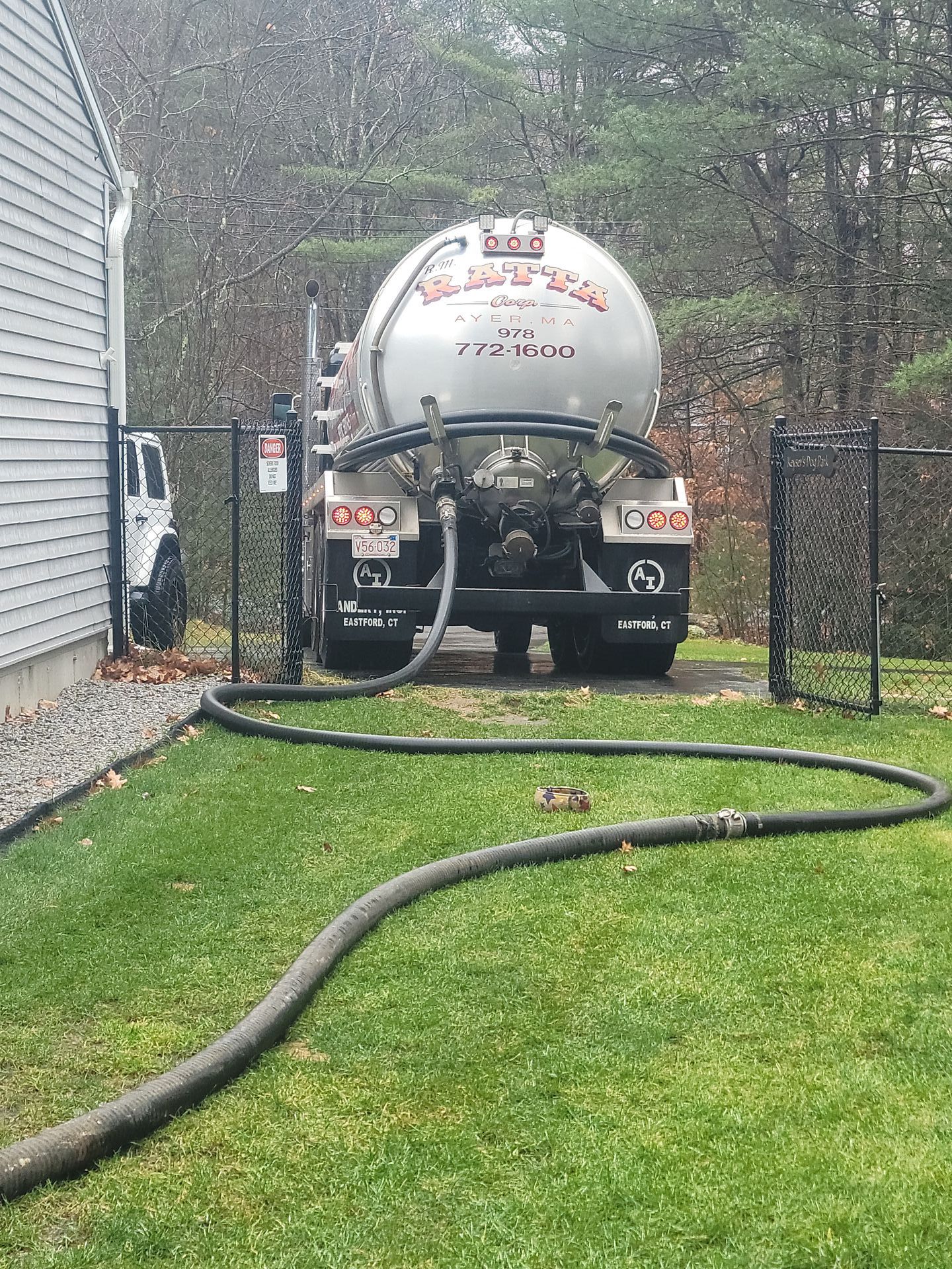 A septic tank truck servicing a residential property, with a hose stretching across the grass. The truck is silver, and the house is beige.