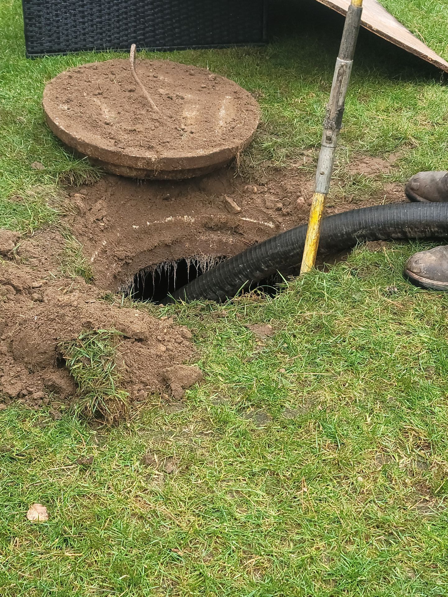 A circular hole in a grassy yard with a manhole cover and black drainage pipe. The soil is dark and the grass is green.