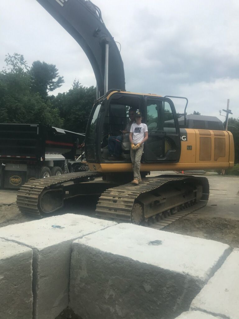 A person stands on a large yellow excavator. A truck sits beside it, and concrete blocks are in the foreground.