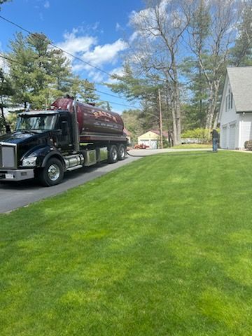 A black septic truck on a green lawn in front of a house, with a man standing near the garage.