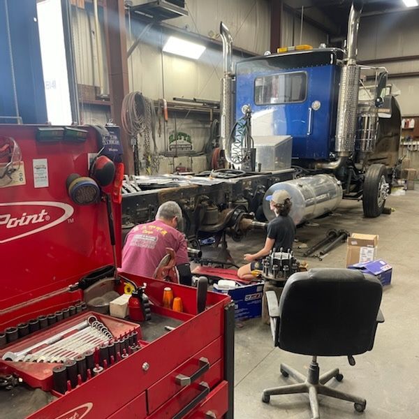 Two mechanics work on a blue semi-truck in a garage, surrounded by tools and parts. A red toolbox sits in the foreground.