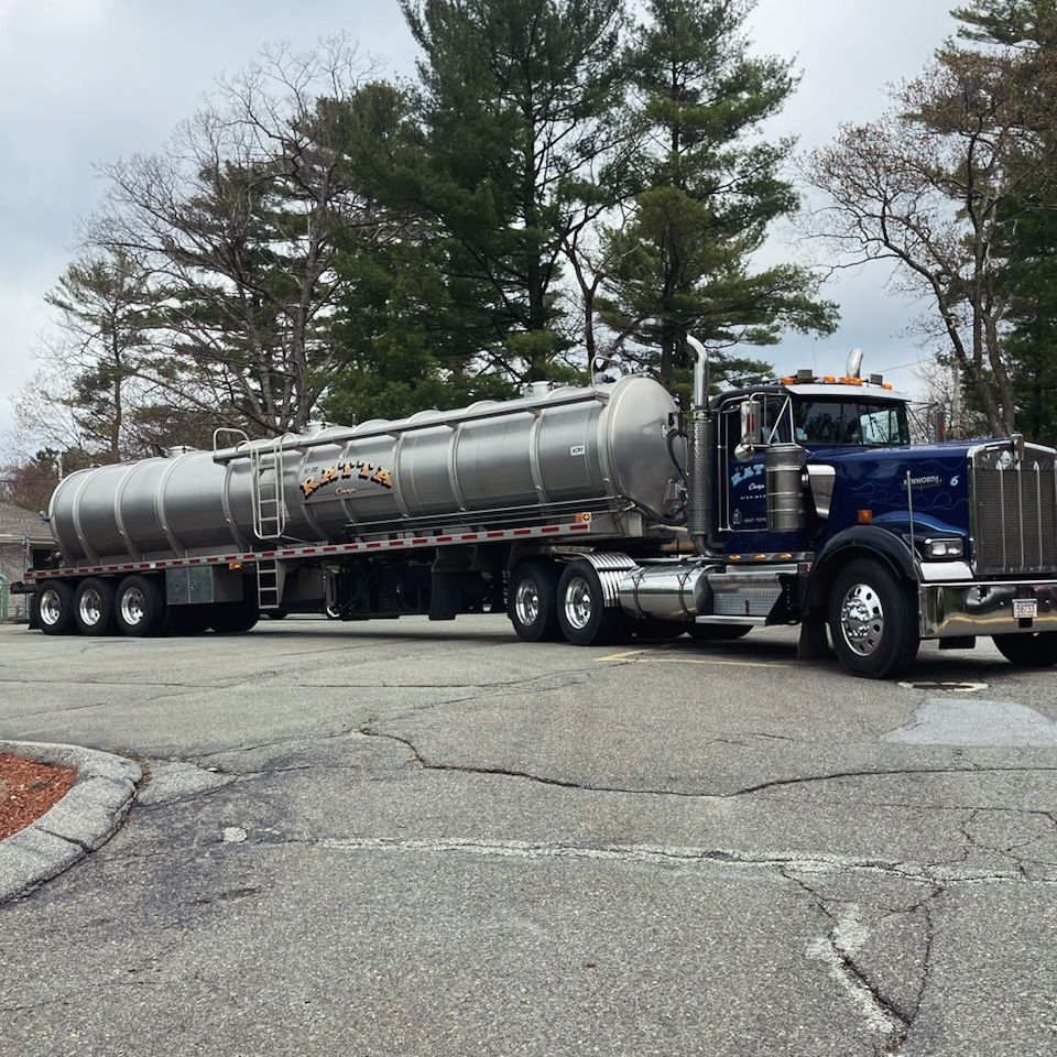 Blue semi-truck pulling two silver tanker trailers on a paved road near trees.