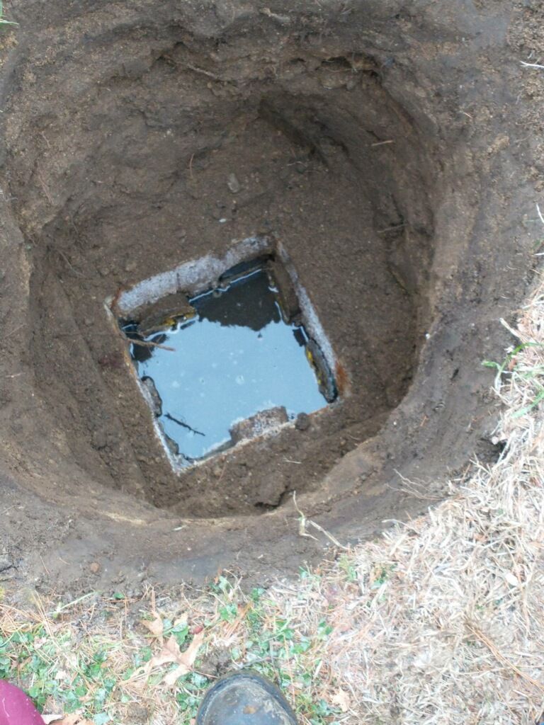 An open square hole in the ground, revealing a metal lid submerged in dark liquid. The surrounding soil is dark brown.