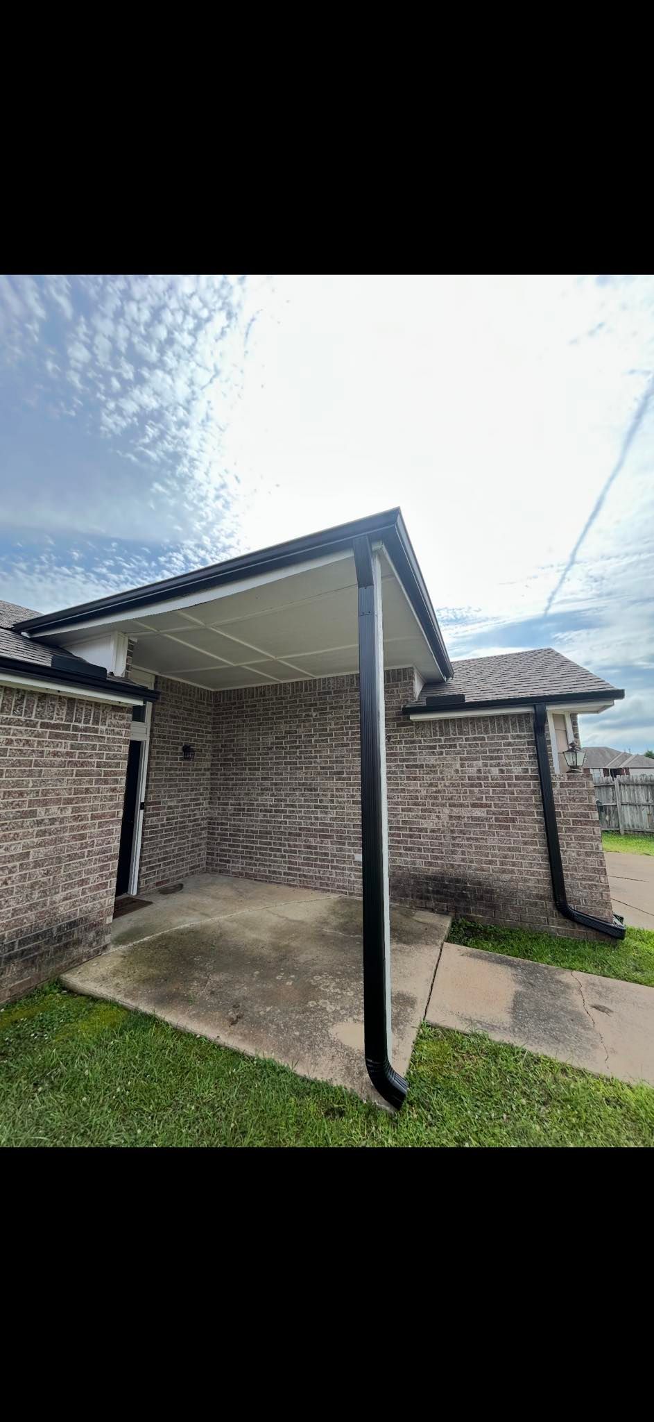 A brick home exterior with a covered patio, a concrete path, a green lawn, and dark-colored gutters under a blue sky.