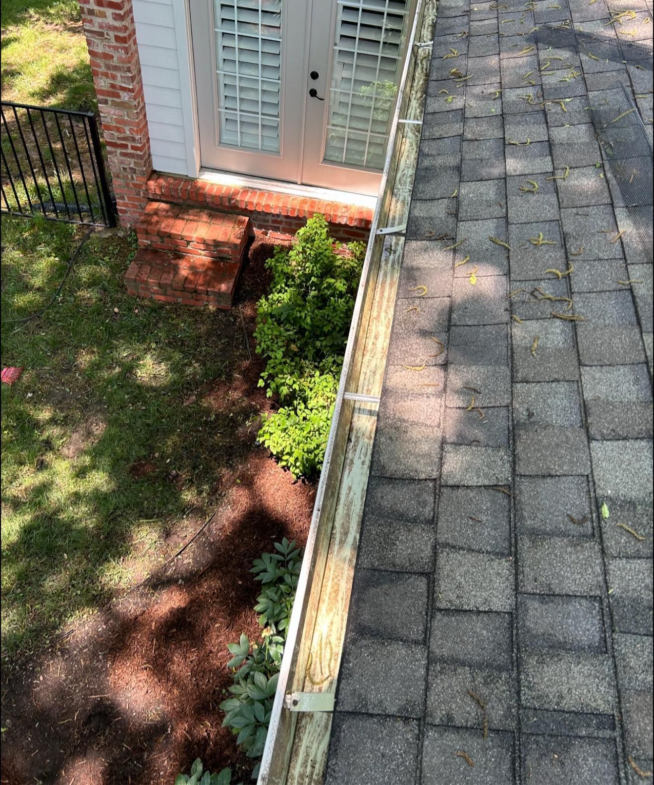 A high-angle view of a grey shingled roof edge with metal gutter clips, overlooking a garden, brick steps, and glass doors.