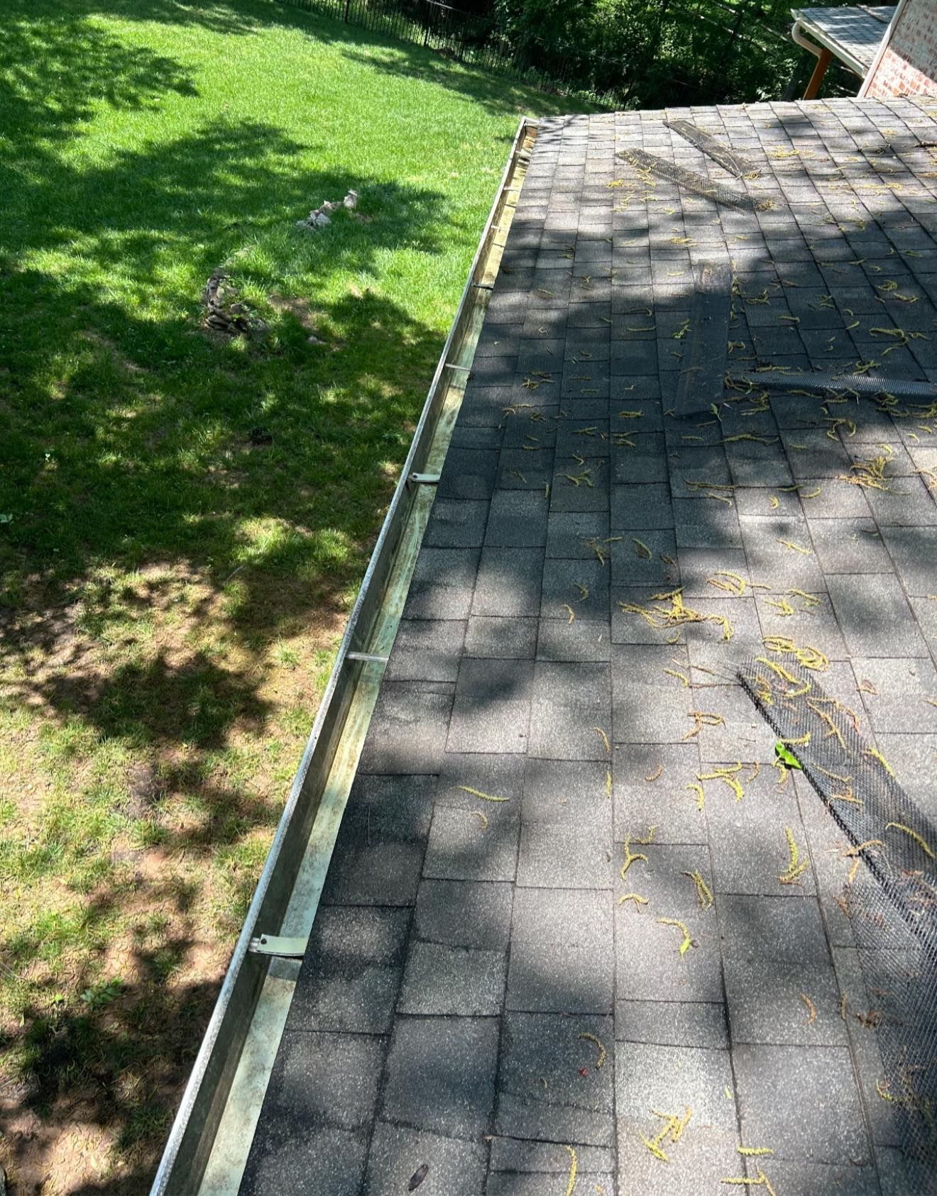 A metal rain gutter attached to the edge of an asphalt shingle roof, overlooking a grassy lawn below.