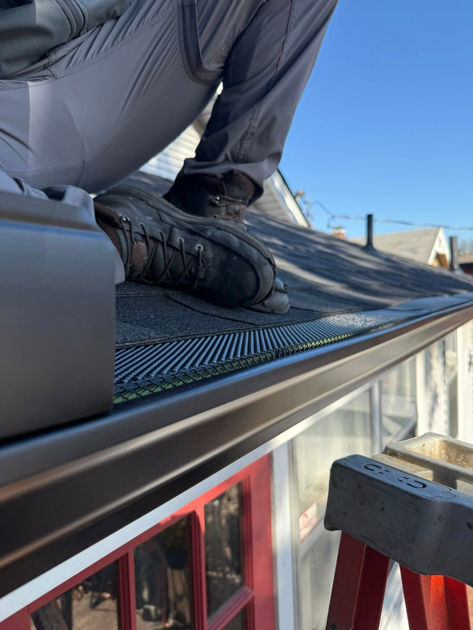 A person in work pants and boots kneels on a roof, installing a dark metal gutter guard system above a red-framed window.