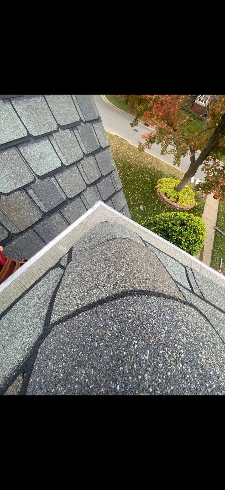 A high-angle view of a grey shingled roof edge meeting a ridge, with a blurred residential yard and tree in the background.