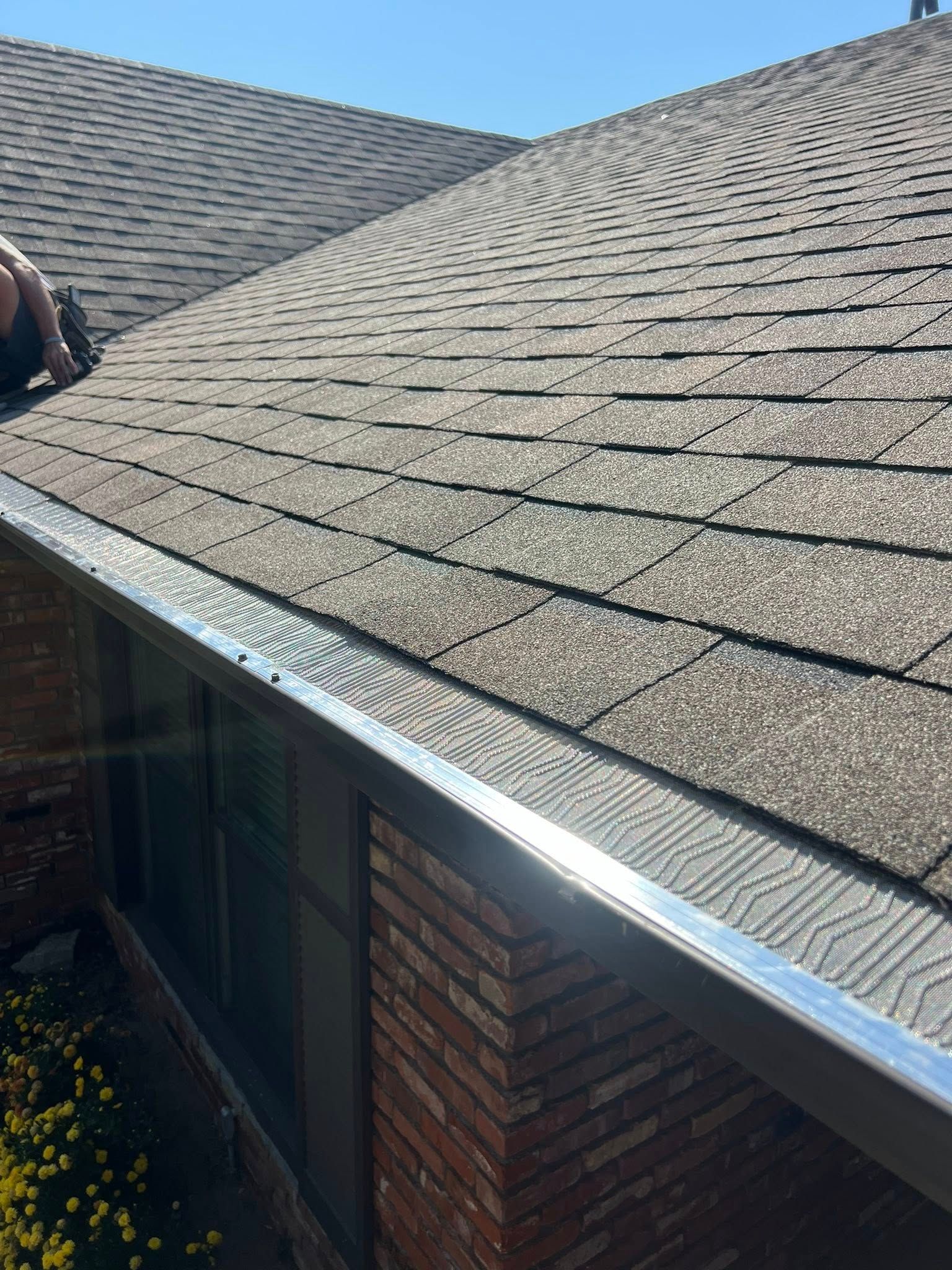 A worker installing metal gutter guards on the edge of a residential shingle roof.