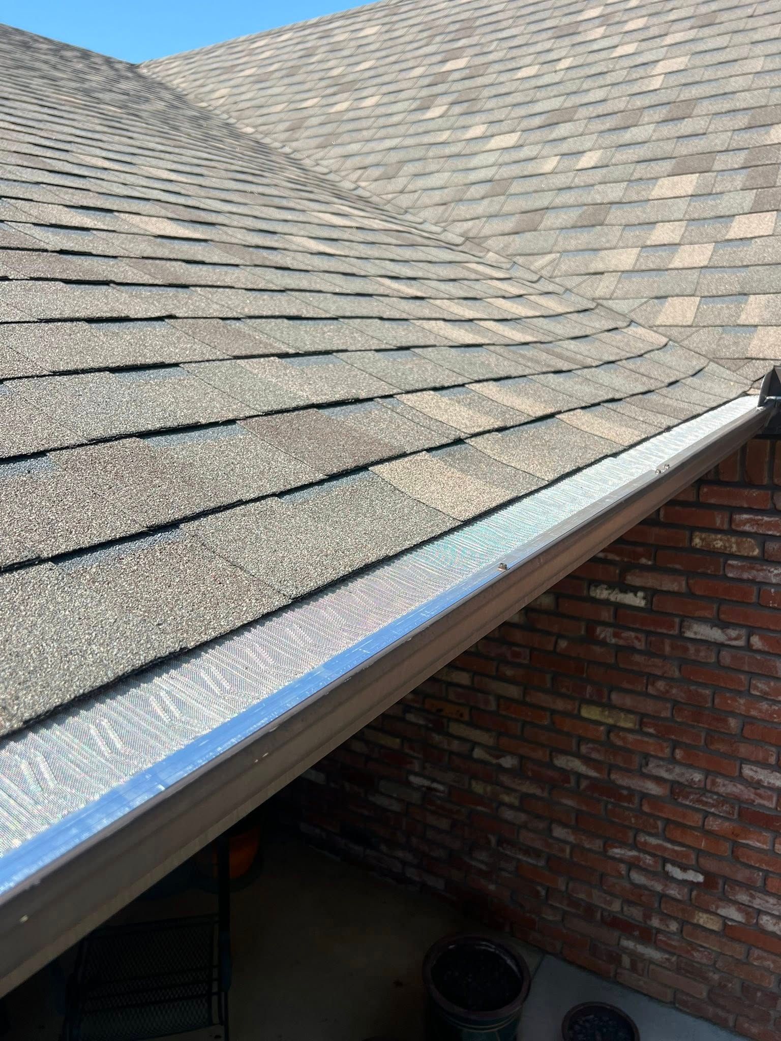 Close-up of a brown shingled roof with a silver metal gutter guard installed along the edge above a brick wall.