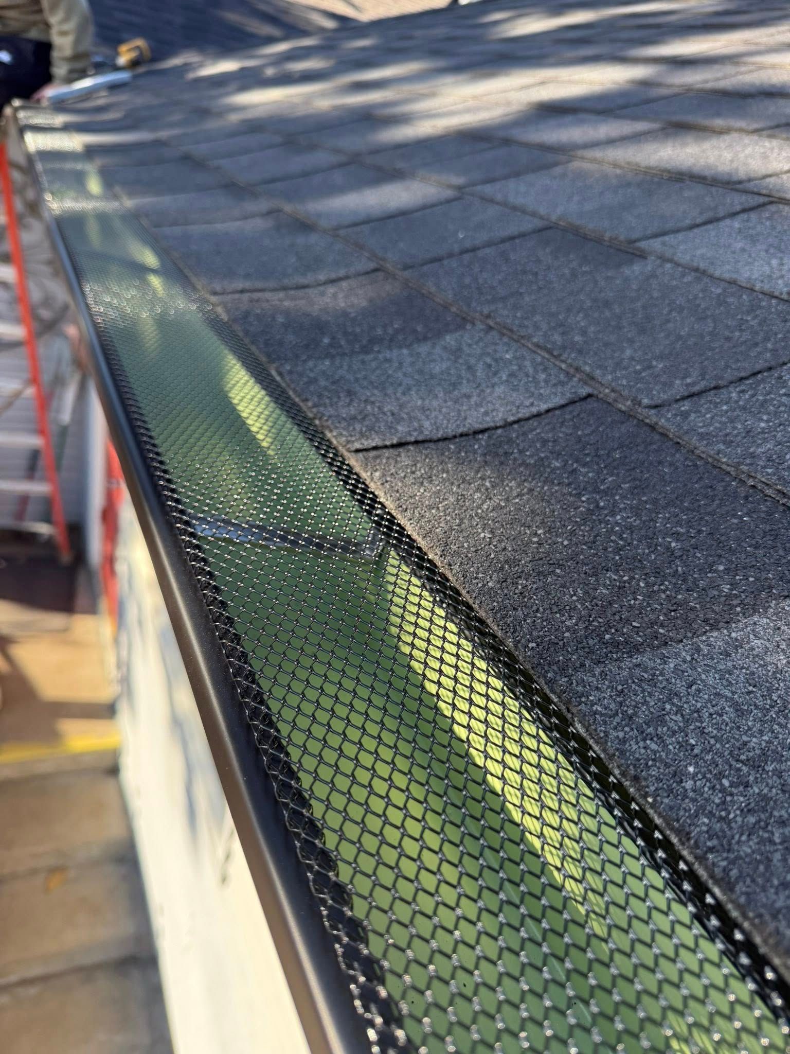 A close-up view of a metal gutter guard installed over a residential gutter along the edge of an asphalt shingle roof.