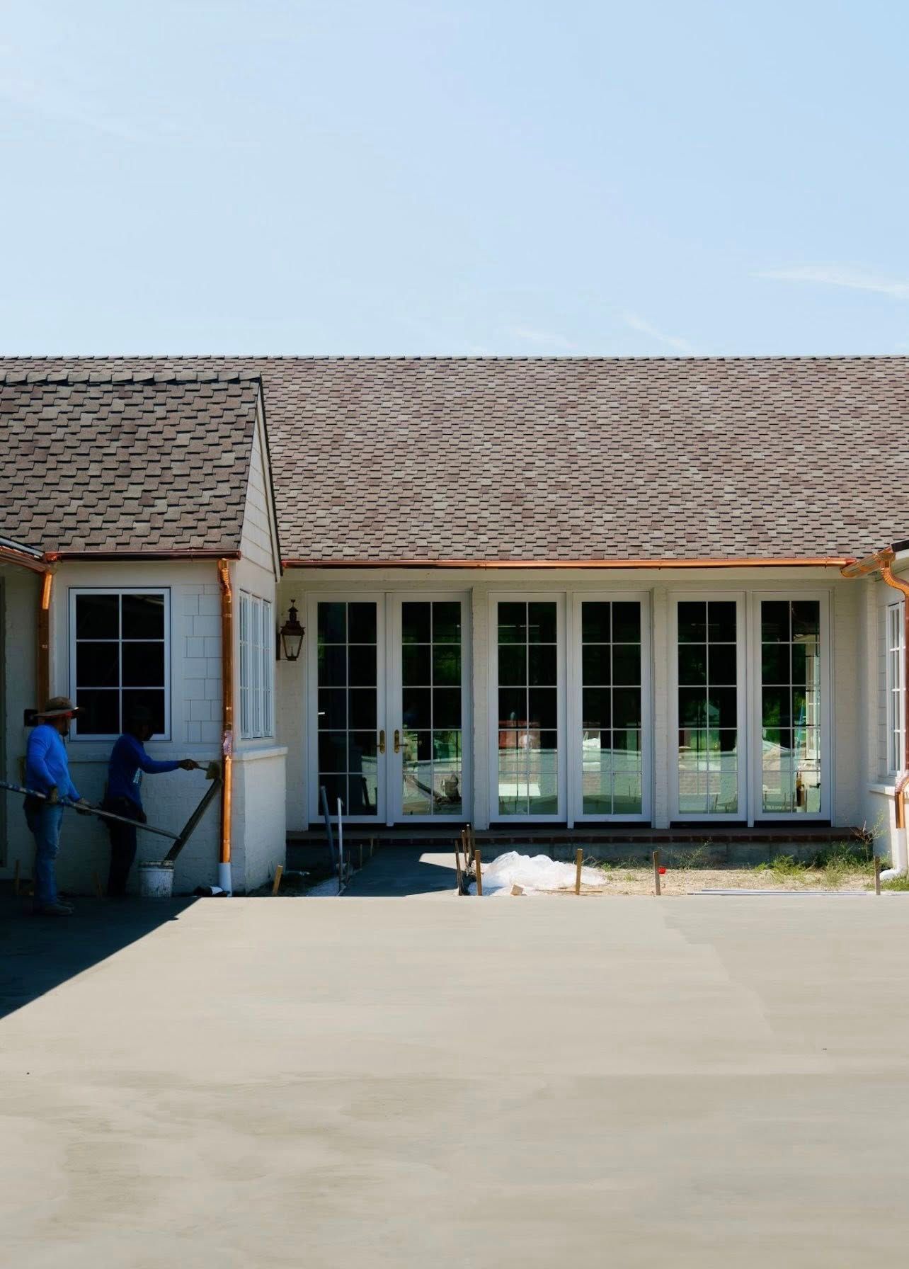 Two workers finish a light-colored concrete patio in front of a house with white-framed glass doors and a shingled roof.