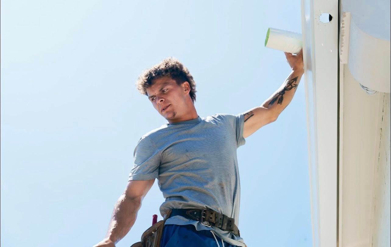 A person wearing a gray t-shirt and work belt reaches up to perform repairs on a white roof structure against a clear sky.