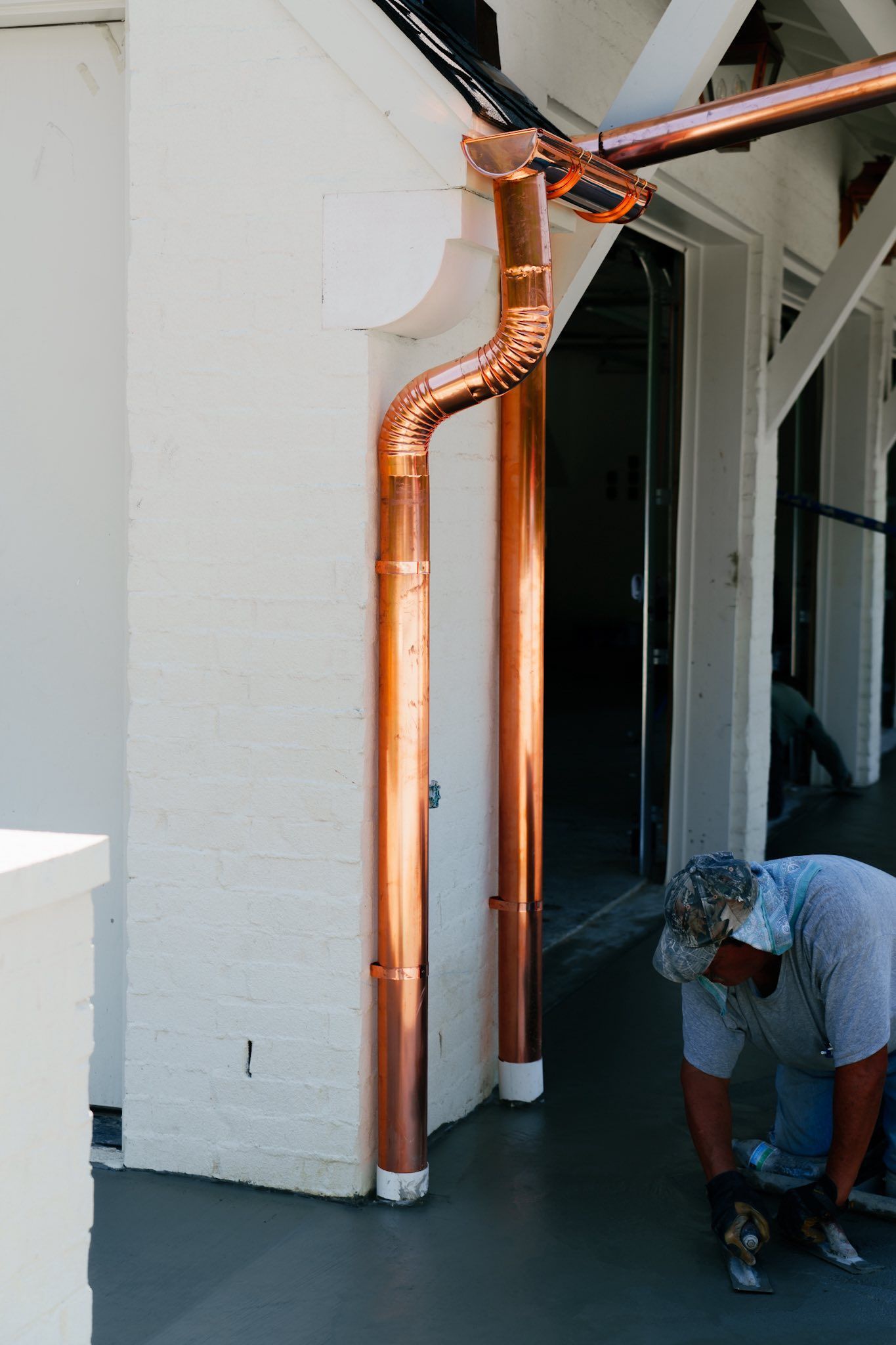 A worker kneels on a gray surface while installing shiny copper downspouts on the corner of a white building.