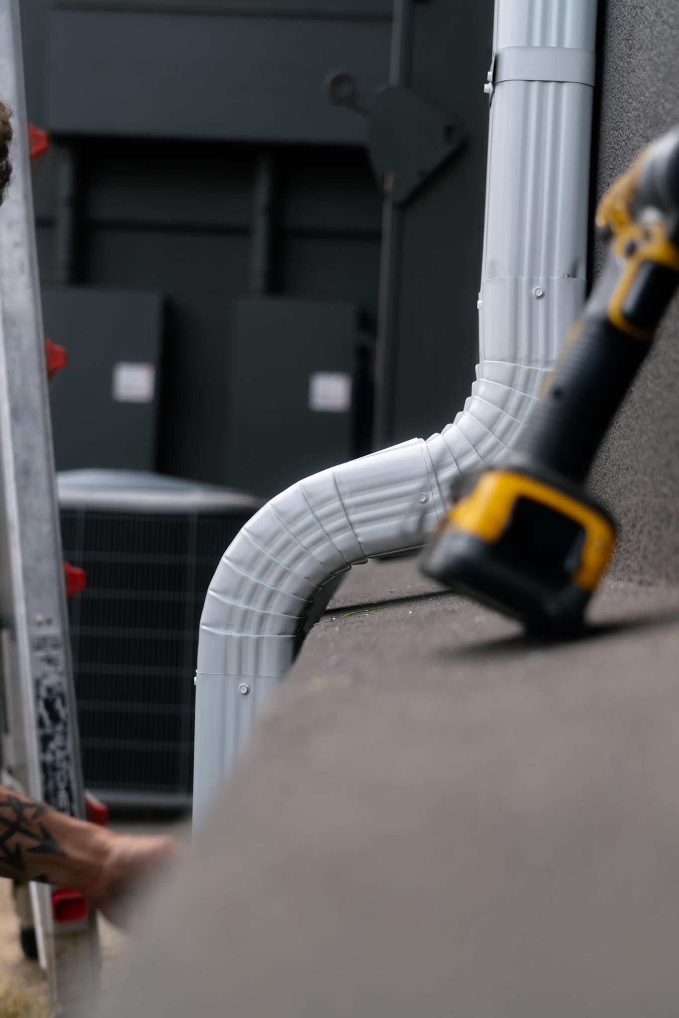 A close-up view of a white gutter downspout being installed on a wall next to a yellow cordless drill.
