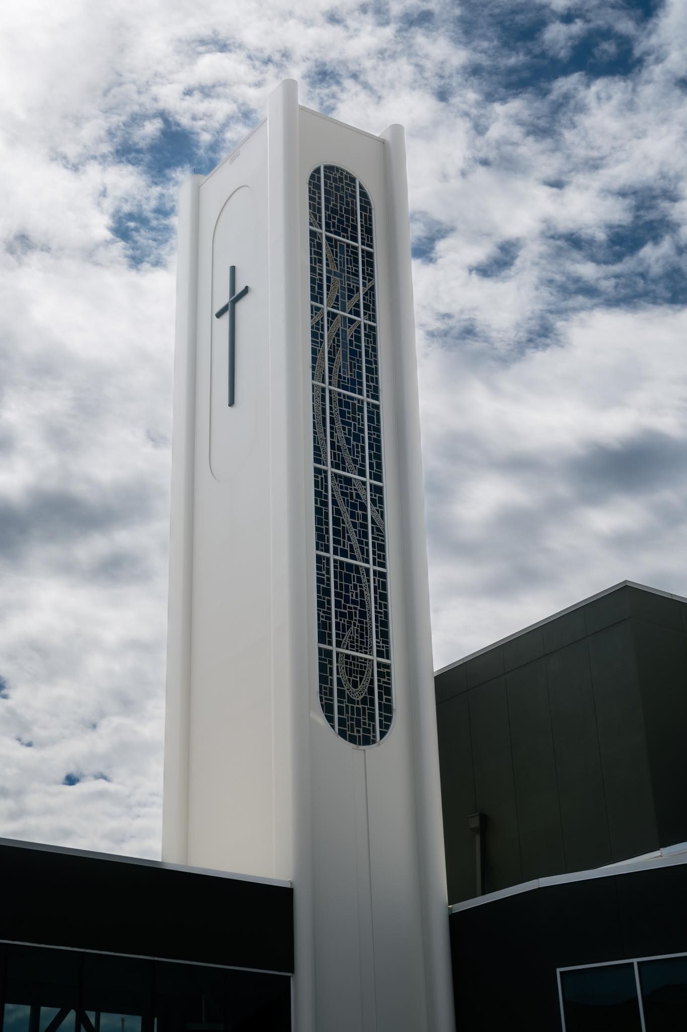 A tall, white church bell tower against a cloudy sky, featuring a dark cross and a long, textured vertical window panel.