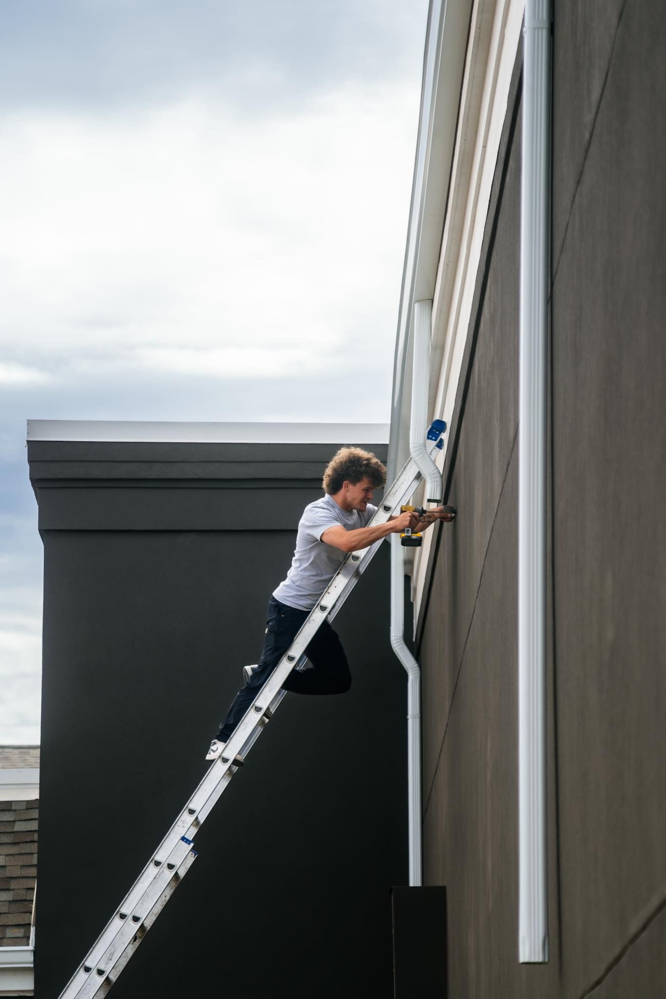 A person in a gray shirt uses a drill while balanced on a ladder leaning against a dark exterior wall.
