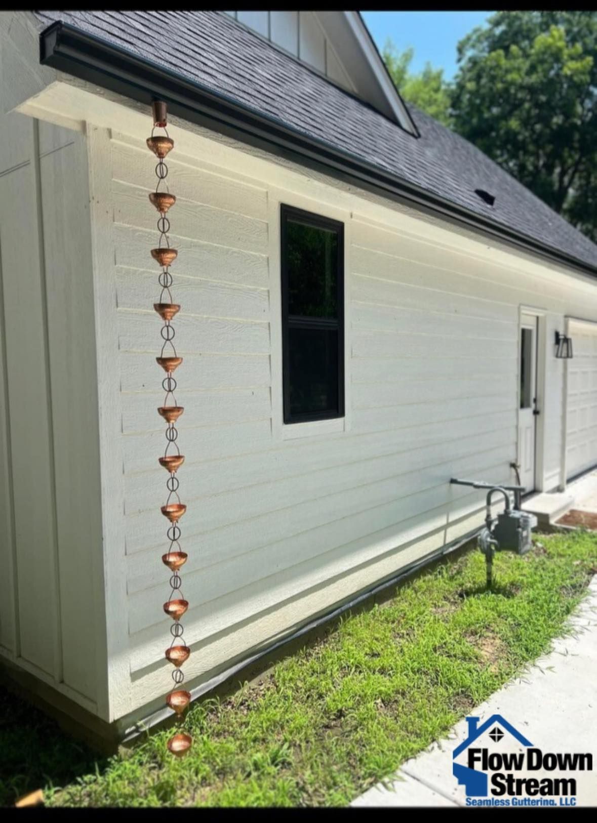 A copper rain chain hangs from the black gutter of a white house, positioned beside a window and a grassy area.
