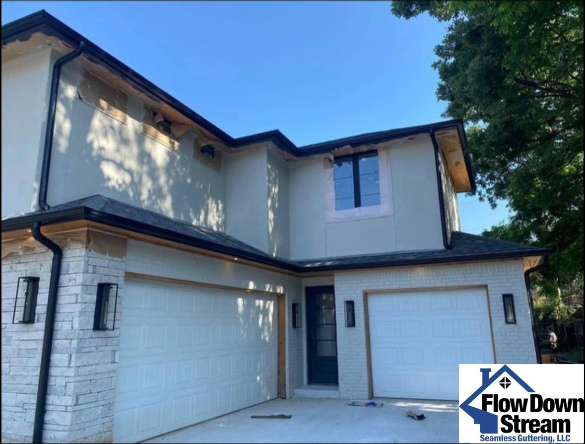 A two-story house exterior with light stucco walls, stone accents, two white garage doors, and black trim under a blue sky.