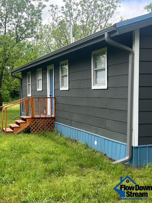 A dark gray mobile home with blue skirting, a wooden porch, and white-trimmed windows set in a grassy yard.