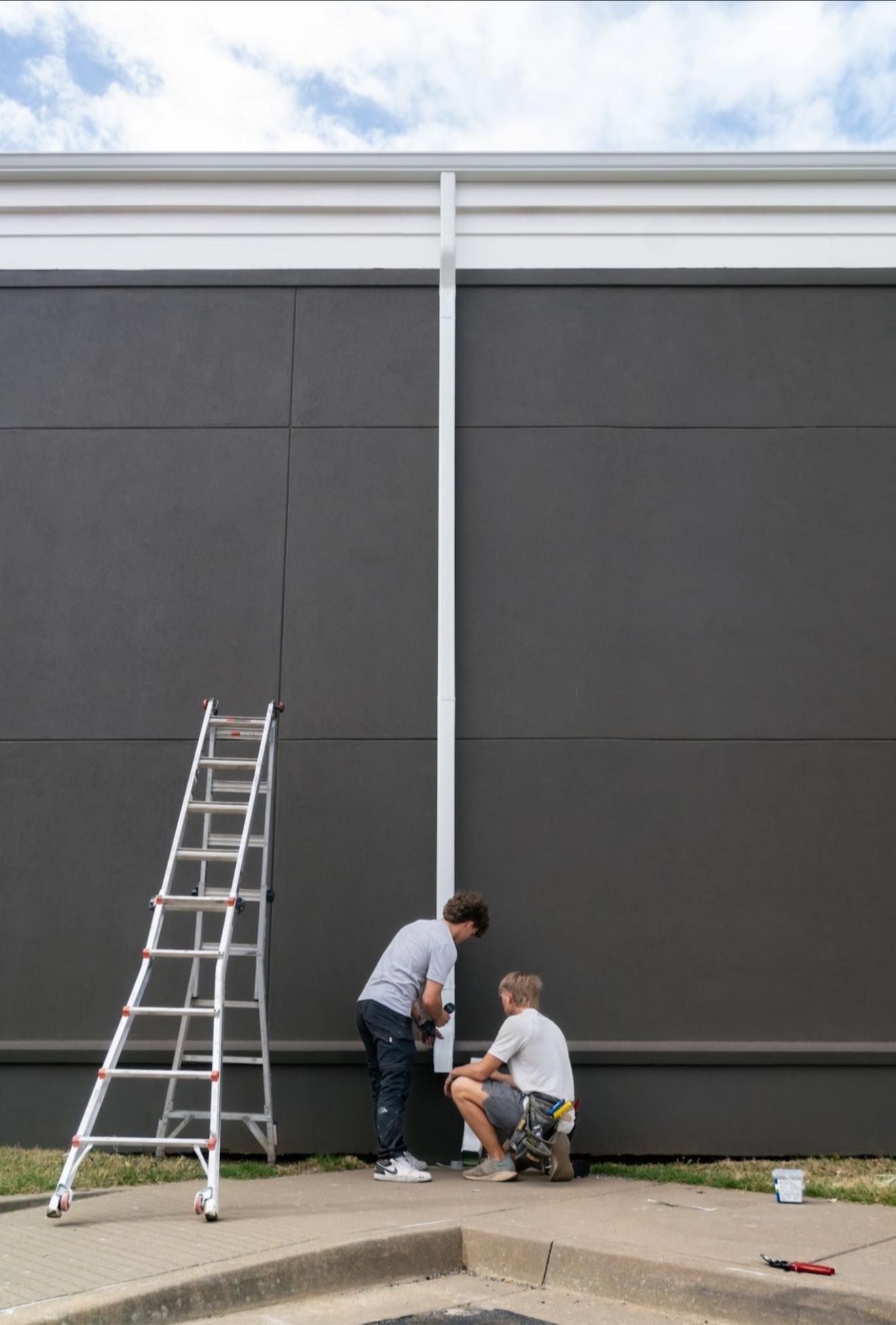 Two people work on a white downspout on a dark gray building wall with a metal ladder standing nearby.