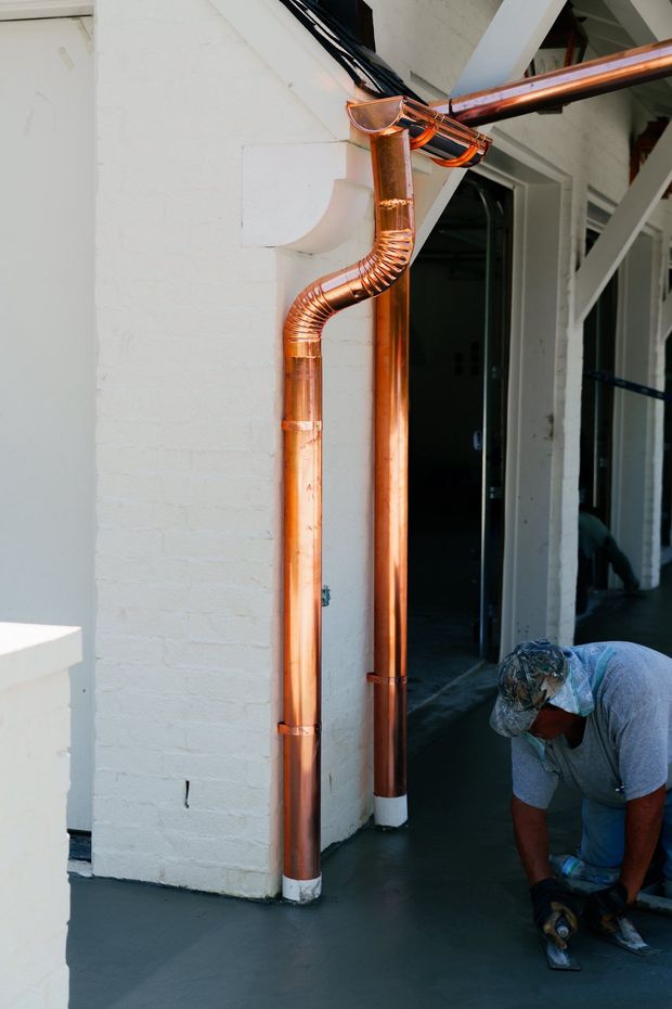 A construction worker installs new copper downspouts against a white brick pillar on a grey patio.