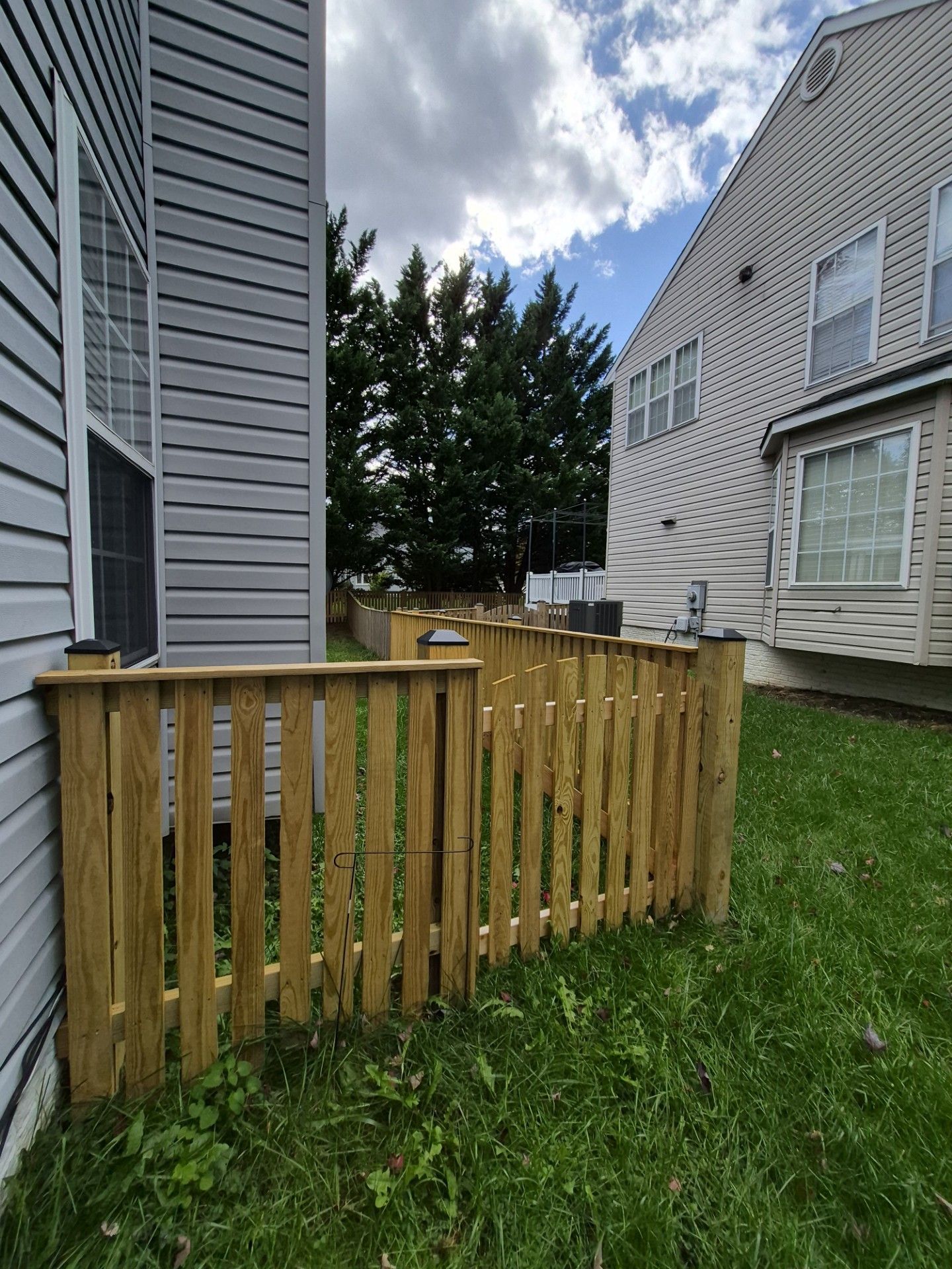 Wooden fence around a grassy area between two houses. The sky is partly cloudy.