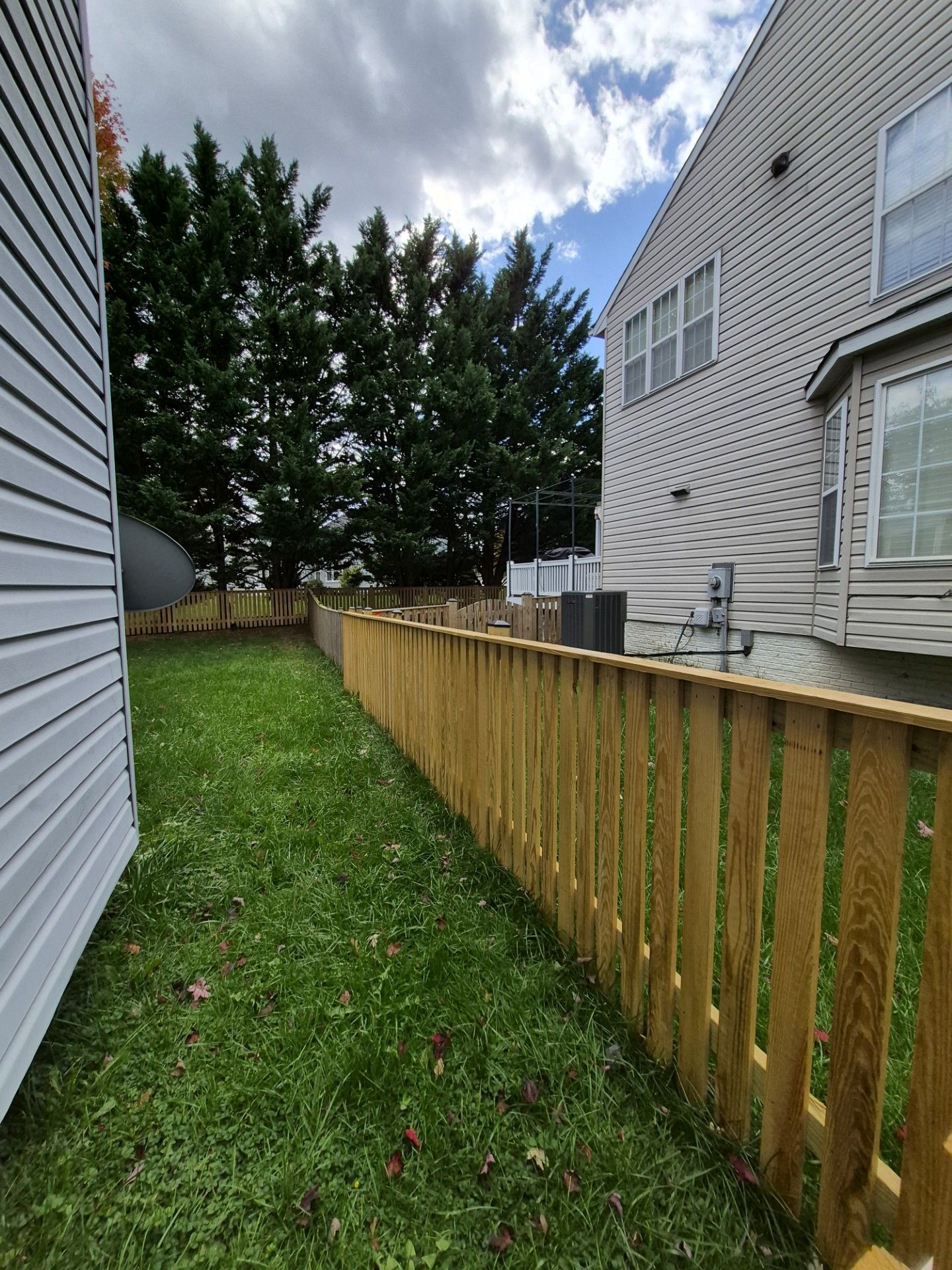 Wooden fence in a grassy yard, between a house with siding and another building, under a cloudy sky.