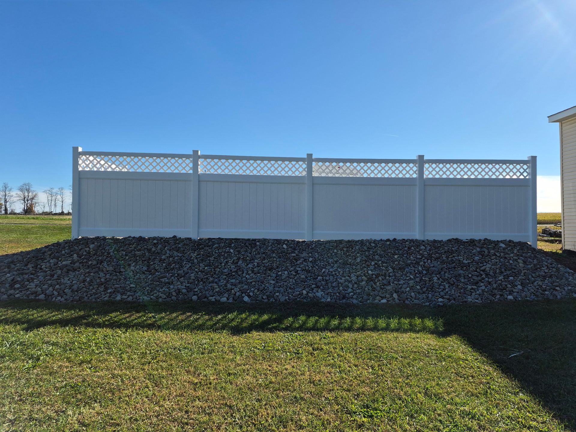 White vinyl fence with lattice top, on a bed of gravel, in a grassy yard under a blue sky.