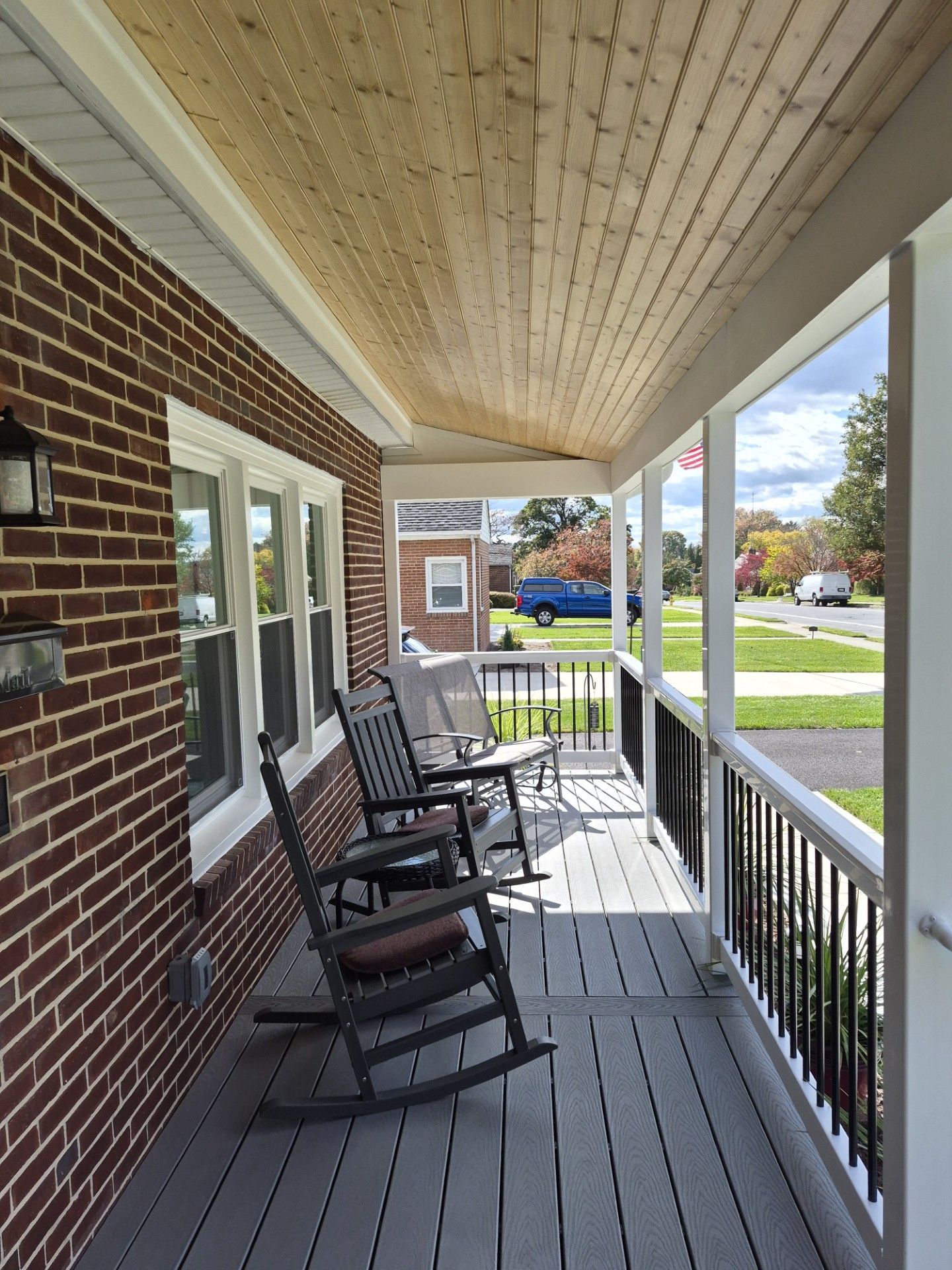 Porch with gray decking, brick wall, and rocking chairs. Wooden ceiling and a view of the street.