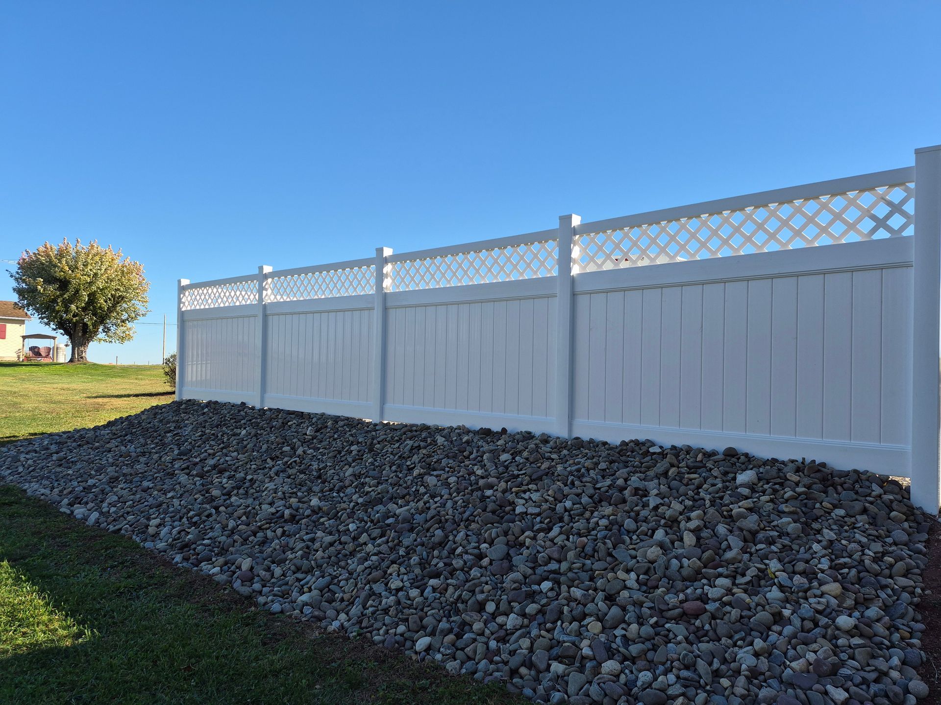 White privacy fence with lattice top, backed by a pile of gray rocks, under a blue sky.